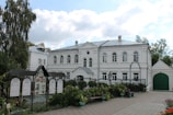 A large, two-story white building with classical architectural features, including arched windows and a triangular pediment. The building is surrounded by lush greenery and a paved courtyard. In the foreground, a decorative fence with display boards is visible, and there are planted pots on either side.