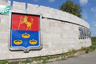 A stone wall with a large colorful shield emblem on the left side. The emblem features a golden lion holding a staff on a red background and three yellow rings on a blue background beneath it. There is greenery and some flowers at the base of the wall. The sky is clear with some clouds and a tree is visible in the background.