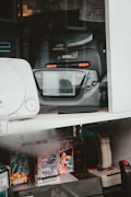 A collection of vintage gaming consoles and accessories is displayed. Prominently featured is a black Sega Game Gear, positioned beside other retro gaming products such as a joystick and video game cartridges. The display includes various models and game titles, suggesting a focus on retro gaming nostalgia.