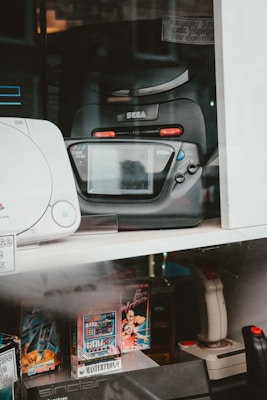 A collection of vintage gaming consoles and accessories is displayed. Prominently featured is a black Sega Game Gear, positioned beside other retro gaming products such as a joystick and video game cartridges. The display includes various models and game titles, suggesting a focus on retro gaming nostalgia.