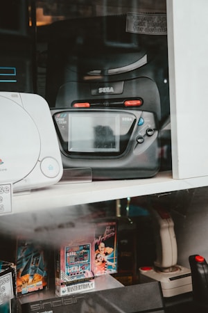 A collection of vintage gaming consoles and accessories is displayed. Prominently featured is a black Sega Game Gear, positioned beside other retro gaming products such as a joystick and video game cartridges. The display includes various models and game titles, suggesting a focus on retro gaming nostalgia.