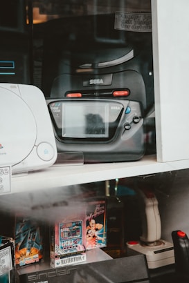 A collection of vintage gaming consoles and accessories is displayed. Prominently featured is a black Sega Game Gear, positioned beside other retro gaming products such as a joystick and video game cartridges. The display includes various models and game titles, suggesting a focus on retro gaming nostalgia.