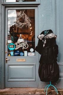 A decorated storefront door with a Christmas wreath and a 'Merry Christmas' sign. There are various stickers and signs on the glass, including 'Shop Local' and payment methods like Visa. To the right of the door, there is a mannequin wearing a black dress and a fur-lined coat.