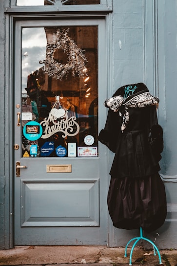 A decorated storefront door with a Christmas wreath and a 'Merry Christmas' sign. There are various stickers and signs on the glass, including 'Shop Local' and payment methods like Visa. To the right of the door, there is a mannequin wearing a black dress and a fur-lined coat.