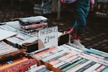 A street market table displaying paperback books for sale at £1 each. The table is filled with various books and DVDs, and a person wearing pink socks and white shoes is visible in the background.