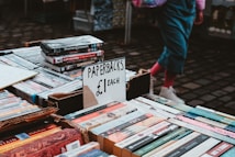 A street market table displaying paperback books for sale at &pound;1 each. The table is filled with various books and DVDs, and a person wearing pink socks and white shoes is visible in the background.