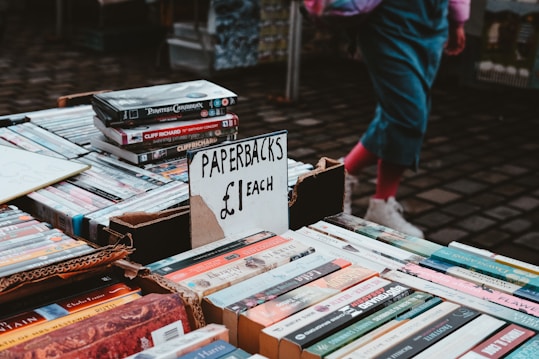 A street market table displaying paperback books for sale at £1 each. The table is filled with various books and DVDs, and a person wearing pink socks and white shoes is visible in the background.