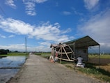 A rural scene with a small structure under construction on the right side of a paved road. Fields extend on either side, with water irrigating the left field. A clear blue sky with scattered clouds creates a bright and open atmosphere. A person sits on a bag at the side of the structure, and a parked vehicle can be seen further down the road.