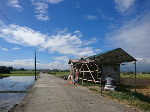 A rural scene with a small structure under construction on the right side of a paved road. Fields extend on either side, with water irrigating the left field. A clear blue sky with scattered clouds creates a bright and open atmosphere. A person sits on a bag at the side of the structure, and a parked vehicle can be seen further down the road.