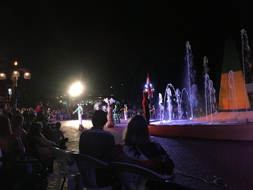 A nighttime outdoor performance featuring dancers in traditional attire. The scene is lit by bright spotlights, with a colorful fountain in the foreground. A crowd of people is sitting and watching the show, creating a lively and festive atmosphere.