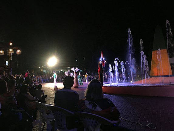 A nighttime outdoor performance featuring dancers in traditional attire. The scene is lit by bright spotlights, with a colorful fountain in the foreground. A crowd of people is sitting and watching the show, creating a lively and festive atmosphere.