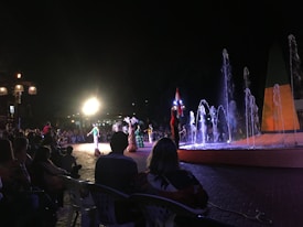 A nighttime outdoor performance featuring dancers in traditional attire. The scene is lit by bright spotlights, with a colorful fountain in the foreground. A crowd of people is sitting and watching the show, creating a lively and festive atmosphere.