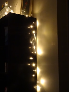 Soft, glowing string lights are adorning the edge of a dark-colored piece of furniture, casting warm reflections on the adjacent wall. A decorative glass bottle sits atop the furniture, adding elegance to the cozy setting.