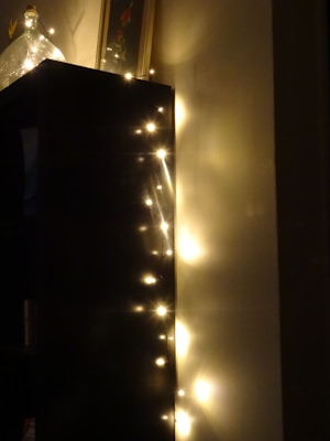 Soft, glowing string lights are adorning the edge of a dark-colored piece of furniture, casting warm reflections on the adjacent wall. A decorative glass bottle sits atop the furniture, adding elegance to the cozy setting.
