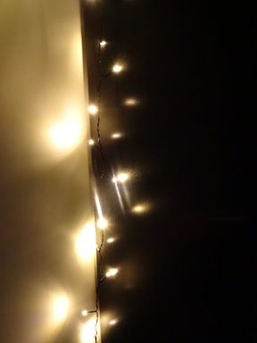 A strand of warm white fairy lights is hung against a dark wall, creating a cozy and intimate ambiance. The lights are small and emit a soft glow, casting gentle reflections on the surface around them.