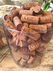 A clear glass container is filled with numerous wine corks, each with unique markings and branding. The corks are densely packed and show a variety of shades from light beige to dark brown. The background includes a blurred wall with natural light and a hint of greenery.