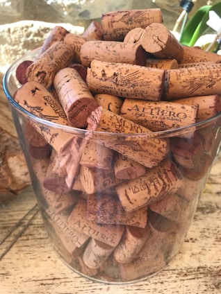 A clear glass container is filled with numerous wine corks, each with unique markings and branding. The corks are densely packed and show a variety of shades from light beige to dark brown. The background includes a blurred wall with natural light and a hint of greenery.