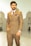 A man wearing a double-breasted brown suit with a white shirt and patterned tie stands confidently. The suit features a pocket square and a chain accessory. His watch is prominently displayed on his wrist. In the background, there is a large mosque-like structure with domes and arches.