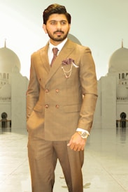 A man wearing a double-breasted brown suit with a white shirt and patterned tie stands confidently. The suit features a pocket square and a chain accessory. His watch is prominently displayed on his wrist. In the background, there is a large mosque-like structure with domes and arches.