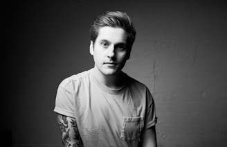 A black and white portrait of a person with short hair and a neutral expression. They are wearing a light-colored t-shirt with rolled-up sleeves, revealing tattooed forearms. The background is a plain wall, providing a minimalistic and focused setting.