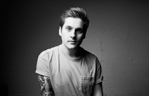 A black and white portrait of a person with short hair and a neutral expression. They are wearing a light-colored t-shirt with rolled-up sleeves, revealing tattooed forearms. The background is a plain wall, providing a minimalistic and focused setting.