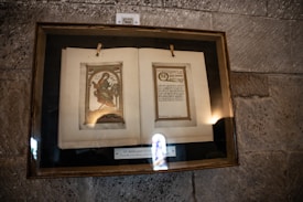 An illuminated manuscript page displayed in a glass frame against a stone wall. The left page features an intricate illustration of a religious figure with vibrant colors, while the right page contains Latin text surrounded by a decorative border. Below the manuscript, a plaque reads 'St. Margaret's Gospel'.