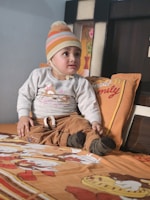 A young child is seated on a bed, wearing a knitted beanie with stripes and casual, comfortable clothing. The bedspread features colorful cartoon characters. A pillow with text and a decorative backdrop with floral designs are visible.