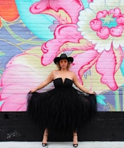 A person stands confidently against a vividly painted brick wall, wearing a black strapless dress with a voluminous tulle skirt. They accessorize with a wide-brimmed black hat and orange-tinted sunglasses. The mural behind them features large, abstract floral designs in bright colors such as pink, turquoise, and yellow.
