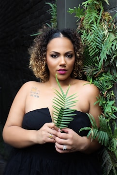 A woman with curly hair and a nose piercing stands against a dark brick wall, holding a leafy green fern in front of her. She is wearing a strapless black dress and has visible tattoos on her shoulder. The wall is adorned with lush green foliage.