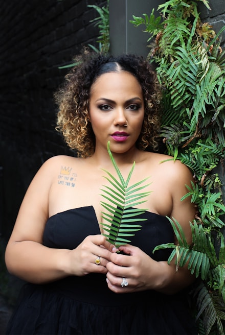 A woman with curly hair and a nose piercing stands against a dark brick wall, holding a leafy green fern in front of her. She is wearing a strapless black dress and has visible tattoos on her shoulder. The wall is adorned with lush green foliage.