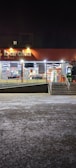 A well-lit storefront of a market at night with large windows displaying goods inside. The store sign reads 'baraka market' and there are stairs leading up to the entrance with an accessible ramp. The exterior lighting illuminates the entrance area, which is otherwise surrounded by darkness.