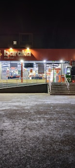A well-lit storefront of a market at night with large windows displaying goods inside. The store sign reads 'baraka market' and there are stairs leading up to the entrance with an accessible ramp. The exterior lighting illuminates the entrance area, which is otherwise surrounded by darkness.