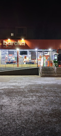 A well-lit storefront of a market at night with large windows displaying goods inside. The store sign reads 'baraka market' and there are stairs leading up to the entrance with an accessible ramp. The exterior lighting illuminates the entrance area, which is otherwise surrounded by darkness.