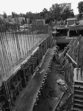 A large construction site with towering metal bars and scaffolding structures. The unfinished concrete foundation is visible, surrounded by various construction materials and tools scattered around the area. Trees and buildings can be seen in the background.