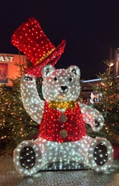 A large festive sculpture of a bear decorated with Christmas lights is illuminated at night. The bear is wearing a red vest and has a matching red top hat. The figure is surrounded by Christmas trees, adding to the holiday atmosphere.