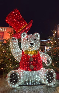 A large festive sculpture of a bear decorated with Christmas lights is illuminated at night. The bear is wearing a red vest and has a matching red top hat. The figure is surrounded by Christmas trees, adding to the holiday atmosphere.
