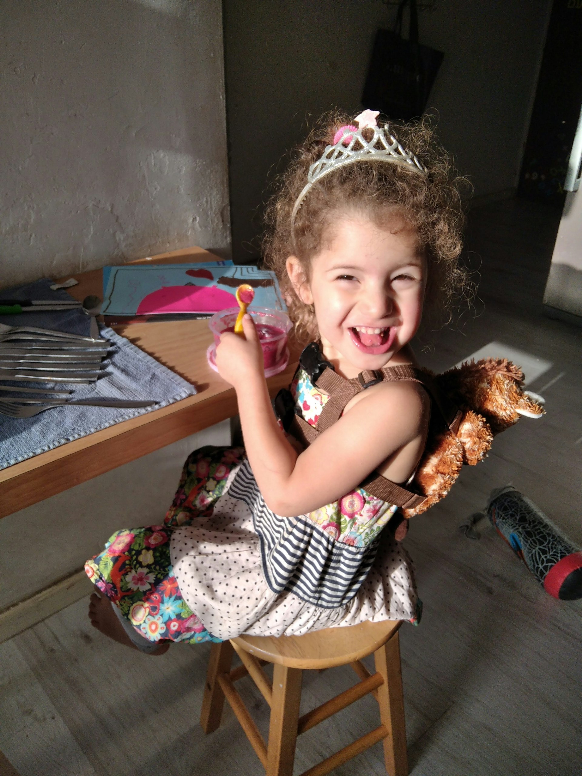 A joyful child with curly hair is sitting on a wooden stool at a table, wearing a decorative tiara and holding a toy spoon in one hand. She is dressed in a colorful, patterned dress, and has a small stuffed animal backpack. Sunlight illuminates her cheerful face, and she is surrounded by various items like a cloth, utensils, a drawing book, and a water bottle on the floor.