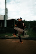 A young baseball player is captured mid-pitch on a pitcher's mound. He is wearing a black jersey with the number one on the back, gray pants, and a blue cap. The background includes a green field, a fence, and a stadium-like structure. The lighting creates a focused atmosphere on the player's intense posture.