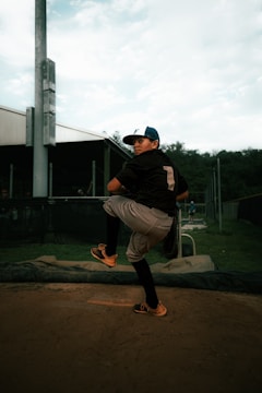 A young baseball player is captured mid-pitch on a pitcher's mound. He is wearing a black jersey with the number one on the back, gray pants, and a blue cap. The background includes a green field, a fence, and a stadium-like structure. The lighting creates a focused atmosphere on the player's intense posture.