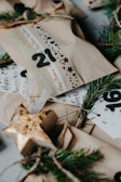 Several wrapped gift packages are decorated with printed ribbons and labels. Sprigs of evergreen are used as embellishments, creating a festive and natural look. The packages are wrapped in plain brown paper, and there are decorative elements like numbers and stars.