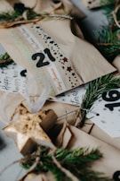 Several wrapped gift packages are decorated with printed ribbons and labels. Sprigs of evergreen are used as embellishments, creating a festive and natural look. The packages are wrapped in plain brown paper, and there are decorative elements like numbers and stars.