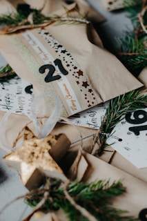 Several wrapped gift packages are decorated with printed ribbons and labels. Sprigs of evergreen are used as embellishments, creating a festive and natural look. The packages are wrapped in plain brown paper, and there are decorative elements like numbers and stars.
