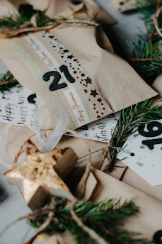 Several wrapped gift packages are decorated with printed ribbons and labels. Sprigs of evergreen are used as embellishments, creating a festive and natural look. The packages are wrapped in plain brown paper, and there are decorative elements like numbers and stars.