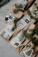 Brown paper bags adorned with festive numbers and decorative foliage, possibly for an advent calendar. Each bag features holiday-themed designs and handwritten messages in a cozy, rustic style. Sprigs of evergreen branch are tied with twine, enhancing the Christmas ambiance.
