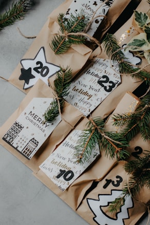 Brown paper bags adorned with festive numbers and decorative foliage, possibly for an advent calendar. Each bag features holiday-themed designs and handwritten messages in a cozy, rustic style. Sprigs of evergreen branch are tied with twine, enhancing the Christmas ambiance.