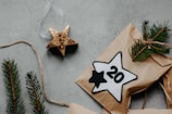 A flat lay featuring a rustic holiday theme. A paper package adorned with pine sprigs is tied with twine. Beside it lies a gold star ornament and a paper star labeled with the number 20. The background is a textured, concrete-like surface.