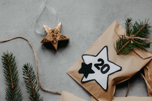 A flat lay featuring a rustic holiday theme. A paper package adorned with pine sprigs is tied with twine. Beside it lies a gold star ornament and a paper star labeled with the number 20. The background is a textured, concrete-like surface.