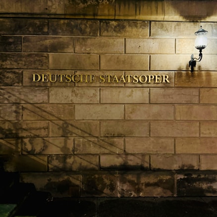 A dimly lit stone wall with the words 'Deutsche Staatsoper' engraved in gold letters near a vintage-style wall lamp on the right-hand side, casting soft light and shadows on the surface.