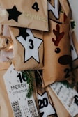 A collection of brown paper bags, each decorated with numbers and festive symbols such as stars and a reindeer face. Some bags have additional decorations like pine branches and glittery tape. A tag labeled 'Merry Christmas' is visible, suggesting an Advent calendar setup.