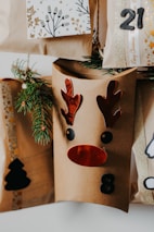 A collection of brown paper gift bags decorated with festive designs, including a reindeer face with red antlers and nose, and numbers like 8 and 21. Sprigs of evergreen are tucked into the bags for decoration. One bag features a black silhouette of a Christmas tree.